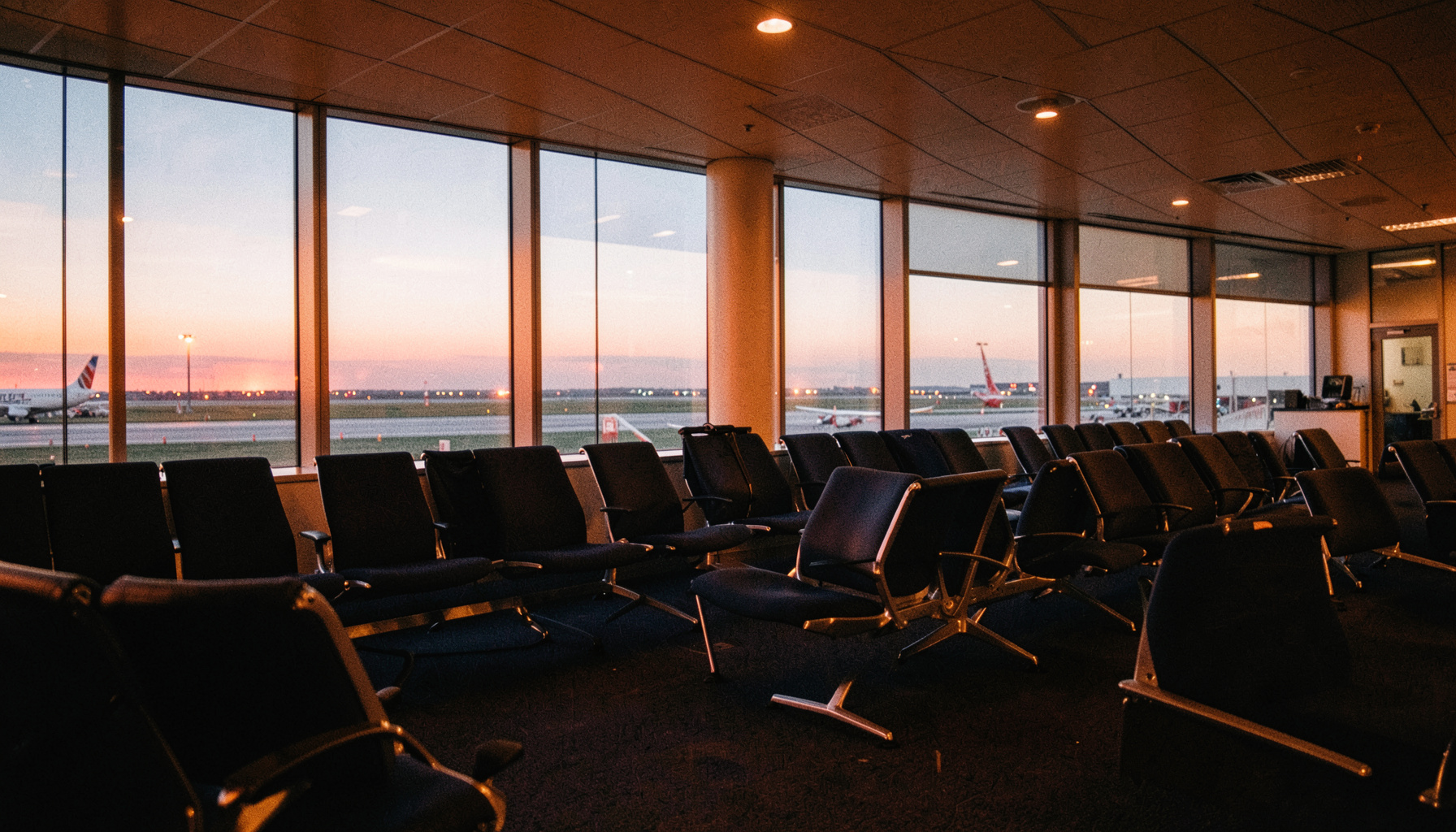 A wide-angle documentary photograph of a quiet airport departure lounge at dusk, warm amber and navy tones, rows of empty seats, large windows showing a runway with soft evening light, calm and anticipatory mood.